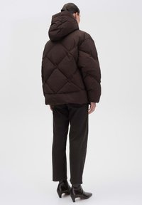 Giubbotto a vento marrone con cappuccio, caratterizzato da un design a trapunta a rombi e una vestibilità rilassata. Indossato con pantaloni neri e stivali con tacco.