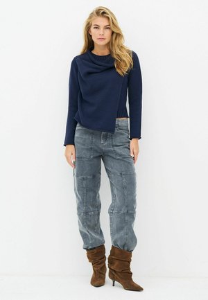 Femme aux longs cheveux blonds portant un pull asymétrique bleu marine, un jean gris patchwork et des bottes marron décontractées à talons, se tenant devant un fond blanc.