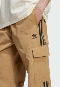 Pantalon cargo beige avec une finition texturée, comportant un logo Adidas, des bandes latérales noires et une poche latérale.
