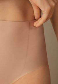 Högmidjade nudefärgade sömlösa underkläder med en slät, elastisk midjeband och mjuk textur, där en hand justerar tyget.
