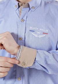 Chemise boutonnée bleu clair avec un motif texturé, featuring des poignets beiges et un accent rayé. Logo brodé sur la poitrine en blanc et bleu.