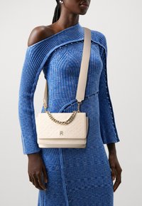 Weißer Handtasche mit strukturiertem, geometrischem Muster und goldener Kettenakzent, quer über ein blaues geripptes Kleid mit asymmetrischem Ausschnitt getragen.