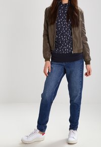 Kvinna klädd i en brun bomberjacka, marinblå mönstrad topp, blå jeans och vita sneakers, stående med avslappnad hållning mot en enfärgad bakgrund.