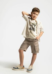 Beżowe szorty cargo, jasna koszula i graficzny t-shirt w delikatnym kremowym odcieniu. Sneakersy z beżowymi, niebieskimi i brązowymi akcentami. Luźny, wygodny strój.