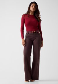 Kobieta stojąca, ubrana w dopasowaną czerwoną bluzkę z długim rękawem i przezroczystym karczkiem oraz ciemnobrązowe, szerokie spodnie z wysokim stanem i dopasowanym paskiem.