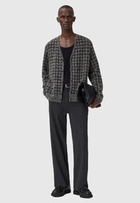 Grå houndstooth cardigan med åpen front, brukt over en svart tanktop. Mørke bukser og svarte sko. Modell som holder en svart clutch.