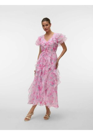 Frau trägt ein fließendes rosa Blumenkleid mit gerafften Ärmeln und Stufen und posiert mit einer Hand in der Hüfte vor einem schlichten weißen Hintergrund.