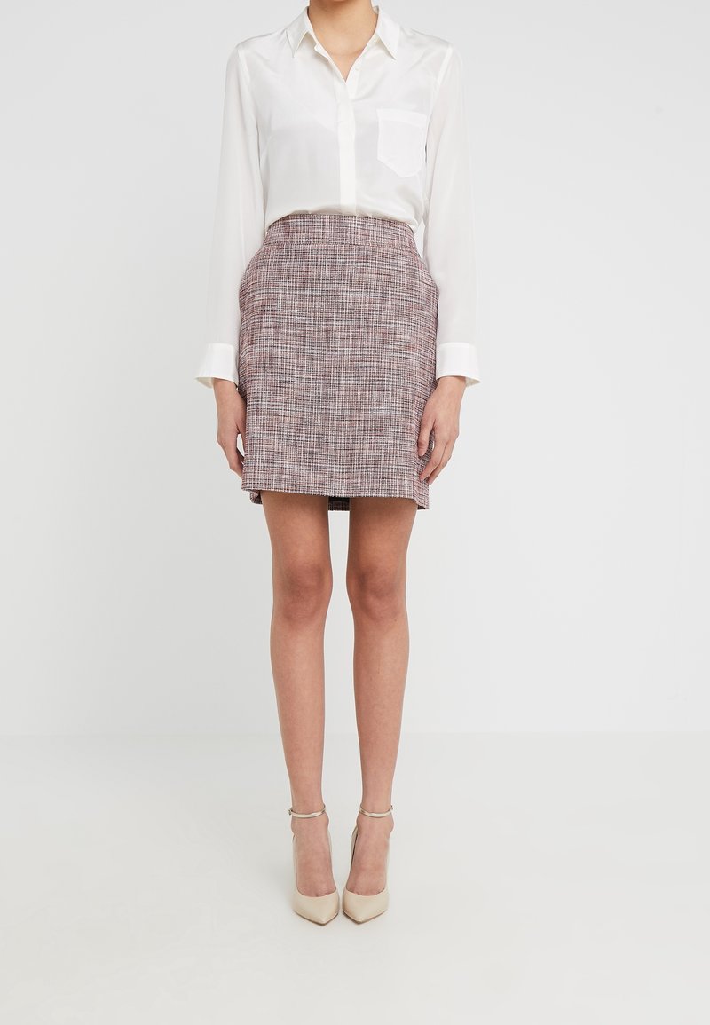 Femme portant un chemisier blanc à manches longues, une jupe à carreaux rose et grise taille haute, et des chaussures à talons beiges, debout sur un fond uni.