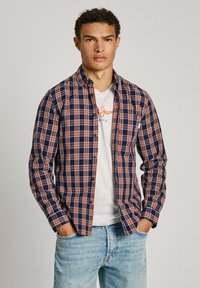 Mornarsko modra in oranžna karirana srajca z dolgimi rokavi, z gumbnim zapenjanjem spredaj in ravnim ovratnikom, oblečena čez svetlo sivo majico. Svetlo modri jeans.