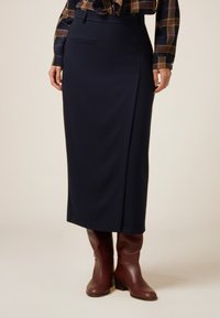 Marineblaue Midi-Rock aus glattem Stoff, mit einem Seitenschlitz, vordere Tasche und strukturiertem Bund. Kombiniert mit braunen Stiefeletten.