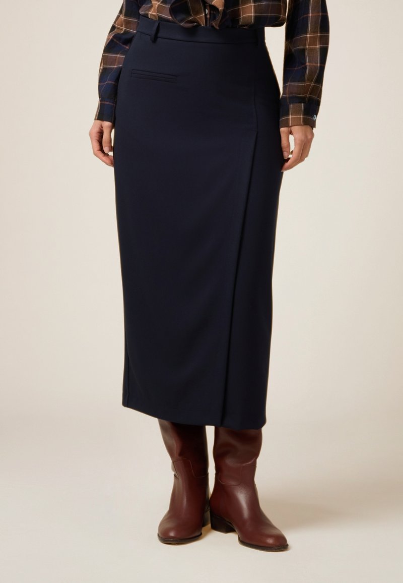 Marineblaue Midi-Rock aus glattem Stoff, mit einem Seitenschlitz, vordere Tasche und strukturiertem Bund. Kombiniert mit braunen Stiefeletten.