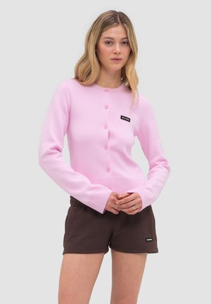 Kobieta w różowym rozpinanym cardiganie i brązowych szortach, stojąca na jednolitym jasnoszarym tle.