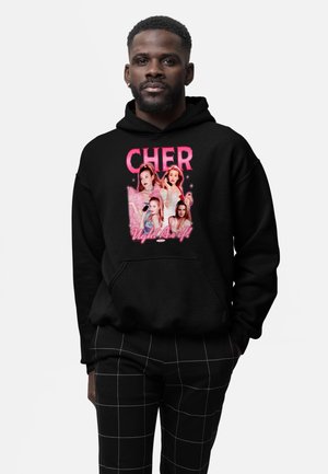 Mand iført sort hættetrøje med pink "CHER" tekst og fire kvindelige portrætter over sætningen "Ugh! As if!", kombineret med sorte ternede bukser.