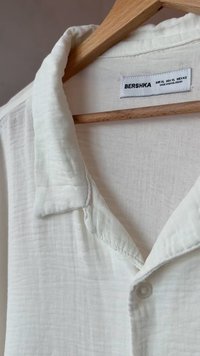 Cămașă albă dintr-un material texturat, cu guler, închidere cu un singur buton și etichetă care afișează marca "BERSHKA" și detalii despre mărime.