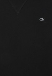 Melnā V veida kakla džemperis, izgatavots no mīksta auduma, ar nelielu baltu izšūtu logo "CK" kreisajā pusē. Gluda tekstūra. Klasicisma dizains.