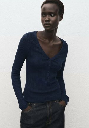 Donna con capelli corti che indossa un cardigan a coste blu navy con scollatura a V e bottoni, abbinato a jeans di denim scuro, in piedi contro un muro bianco testurizzato.