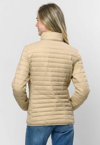 Bežs iešūtā jaka ar horizontālu šuvumu, augstu apkakli un garām piedurknēm. Valkāta kopā ar ziliem džinsiem, parādot pielāgotu siluetu.