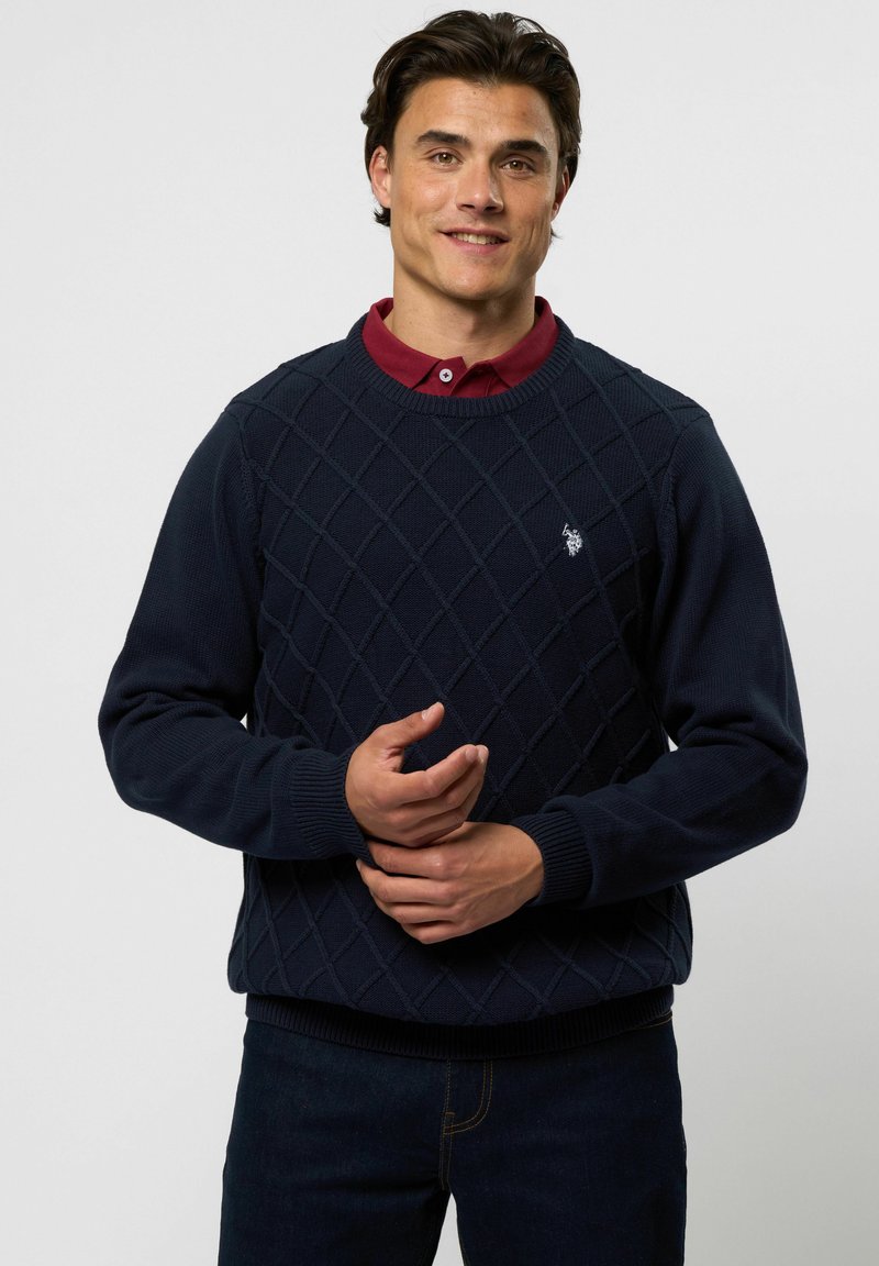 Mann trägt einen marineblauen Pullover mit Diamantmuster über einem kastanienbraunen Hemd mit Kragen, steht mit verschränkten Händen vor hellem Hintergrund.