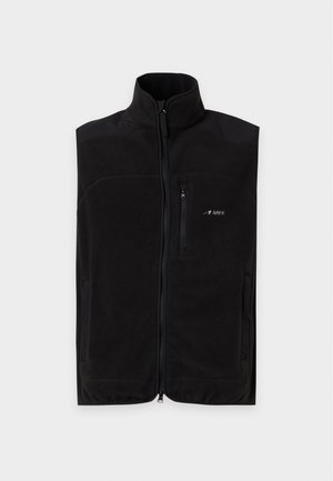 Jeune homme debout face à l’avant, portant un gilet noir sans manches zippé, une chemise noire, un pantalon beige à jambes larges et des chaussures noires avec semelles marron clair.