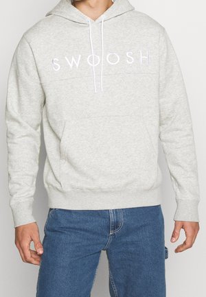 Homme portant un sweat à capuche gris clair avec le texte blanc "SWOOSH" et un jean bleu, debout devant un fond clair uni.