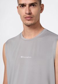 Uomo che indossa una maglietta senza maniche di colore grigio chiaro della Champion, rivolto in avanti su uno sfondo chiaro e semplice.