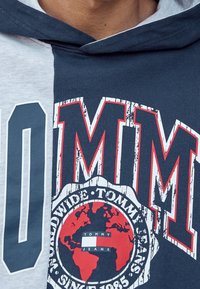 Capuchontrui met een color-block ontwerp in grijs en marineblauw. Voorzien van gedurfde rode en witte grafische tekst en een cirkelvormig logo met een wereldbolontwerp.