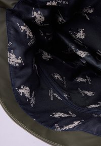 Zielona torba materiałowa z wnętrzem w granatowym kolorze, z powtarzającymi się jasnoszarymi logo U.S. Polo Assn. oraz kieszonką na zamek. Gładka tekstura.