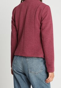 Burgundy texturerad jacka med ribbat mönster, har en avslappnad passform och hög krage, kombinerad med ljusblå denimjeans.