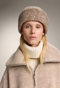 Bežasti pahuljasti šešir i odgovarajući kaput s visokim ovratnikom, u kombinaciji s kremastim rebrastim džemperom s turtleneck izrezom. Mekani materijali i suptilne nijanse.