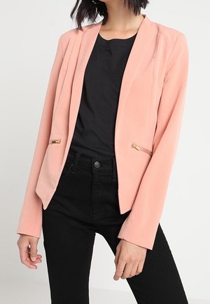 Kvinde iført en lys pink blazer med guld lynlåslommer over en sort skjorte og sorte jeans, stående mod en ensfarvet baggrund.