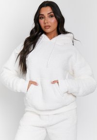 Haut à capuche en polaire blanche et duveteuse avec une poche kangourou ; il possède une capuche ajustable par cordon et des poignets côtelés. La surface texturée offre de la chaleur.