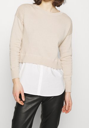 Vrouw draagt een beige gebreide trui over een witte blouse met knoopsluiting en zwarte leren broek, staand tegen een effen achtergrond.