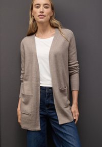 Beige stickad cardigan med öppen framsida, ribbade ärmslut och två framfickor, i kombination med en vit skjorta och mörkblå jeans. Neutral bakgrund.