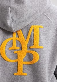Grijze hoodie met grote, gestructureerde gele letters "M," "G," en "P" die elkaar overlappen op de achterkant bij het schoudergebied.
