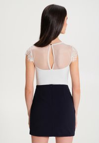 Body bianco con maniche in pizzo trasparente e apertura sul retro, abbinato a una gonna aderente blu navy. Semplice chiusura a bottone al collo.
