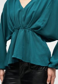 Τιρκουάζ μπλούζα με μακριά μανίκια, σφιγμένη στη μέση και V-λαιμόκοψη, συνδυασμένη με μαύρο δερμάτινο παντελόνι, τα χέρια χαλαρά στο πλάι.