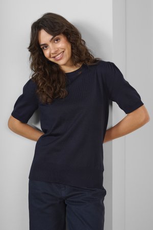 Navy knitted top with short puff sleeves, round neck, and textured fabric; paired with dark pants against a neutral background.