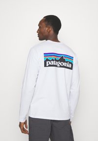 Fehér, hosszú ujjú póló, színes hegyvonulat grafikával, melyen a "patagonia" felirat található a hátsó részen. Az anyag lágy és tartós.