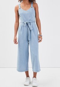 Light blue denim jumpsuit with wide legs, adjustable straps featuring silver hardware, and a tied waist belt. Pairing worn with white sneakers.