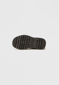 Suela de zapato de goma negra con surcos de tracción profundos y bordes redondeados, que presenta una sección suave en el talón y la marca de la marca.