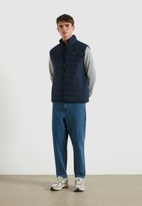 Marinefarbener Steppwesten mit Reißverschluss, seitlichen Taschen und kleinem Logo, getragen über einem grauen Langarmshirt und kombiniert mit blauen Denim-Jeans.