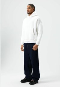 Witte hoodie met capuchon en een buidelzak aan de voorkant, gecombineerd met donkerblauwe jeans met wijde pijpen en zwarte dikke veterschoenen.