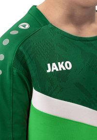 Maglia sportiva verde con tessuto testurizzato, una striscia bianca e logo "JAKO" in bianco. Presenta accenti circolari e un colletto rotondo.