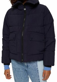 Personen har en sort pufferjakke med frontlynlås, dobbelt klaflommer, lyseblå ribbede sweaterærmer og lyseblå jeans.