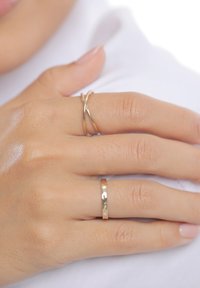 Trys žiedai ant rankos: vienas išlenktas auksinis metalas, vienas lygus auksinis žiedas ir vienas kaltinis auksinis žiedas. Oda šviesiai ruda su natūralia tekstūra.