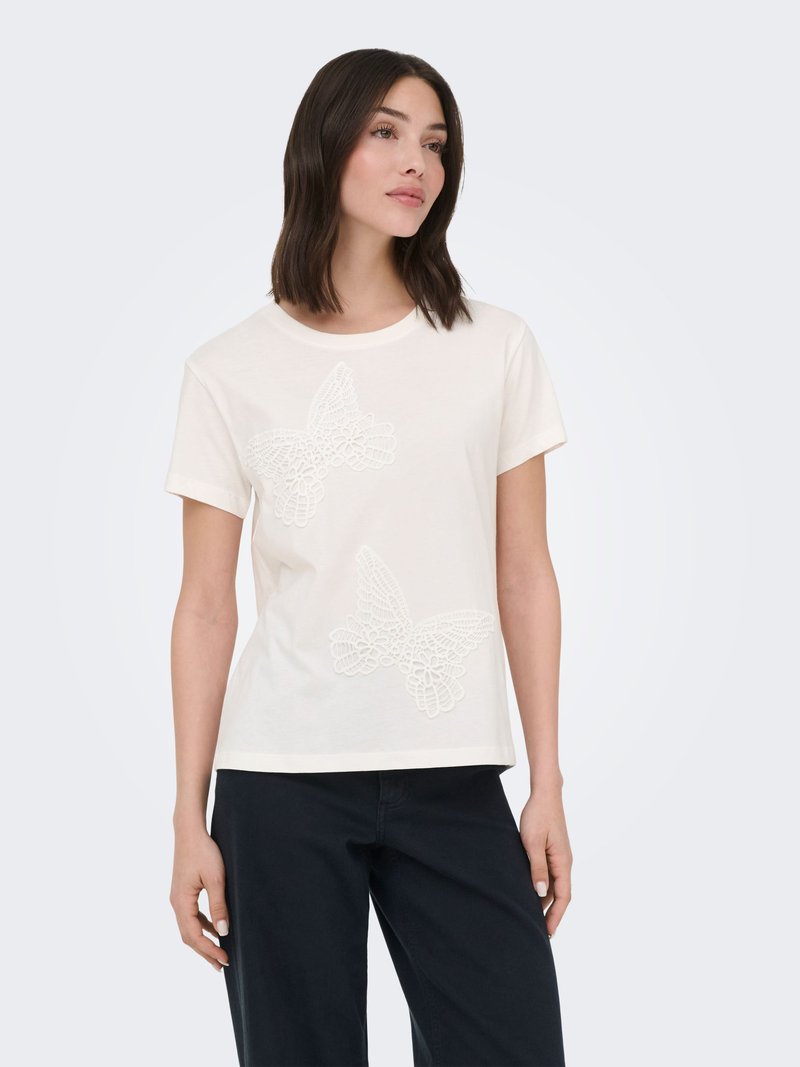 Femme portant une chemise blanche à manches courtes avec des motifs papillons en dentelle sur le devant et un pantalon noir sur un fond uni.