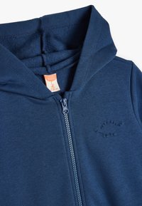 Granatowy baby hoodie wykonany z miękkiej bawełny, z pełnym zamkiem błyskawicznym i teksturowanym logo na klatce piersiowej. Posiada pojemny kaptur.