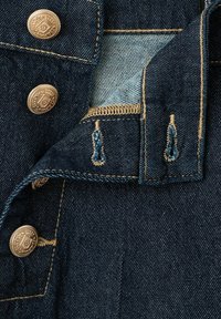 Tmavomodrá džínovina so zlatými gombíkmi a dierkami na gombíky, zobrazujúca detailné šitie a čiastočne otvorenú klopu odhaľujúcu svetlejšiu džínovinu vo vnútri.