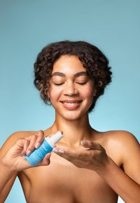 Eine türkisfarbene Hautpflegeflasche mit einem weißen Pumpaufsatz, die in einer Hand gehalten wird und ein klares Serum in die Handfläche der anderen Hand abgibt.