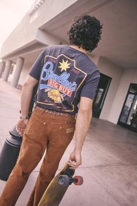 Grafikus póló sötétszürkében, sz�ínes logóval, barna kordnadrággal párosítva. Gördeszkás, aki piros kerekű gördeszkát tart.